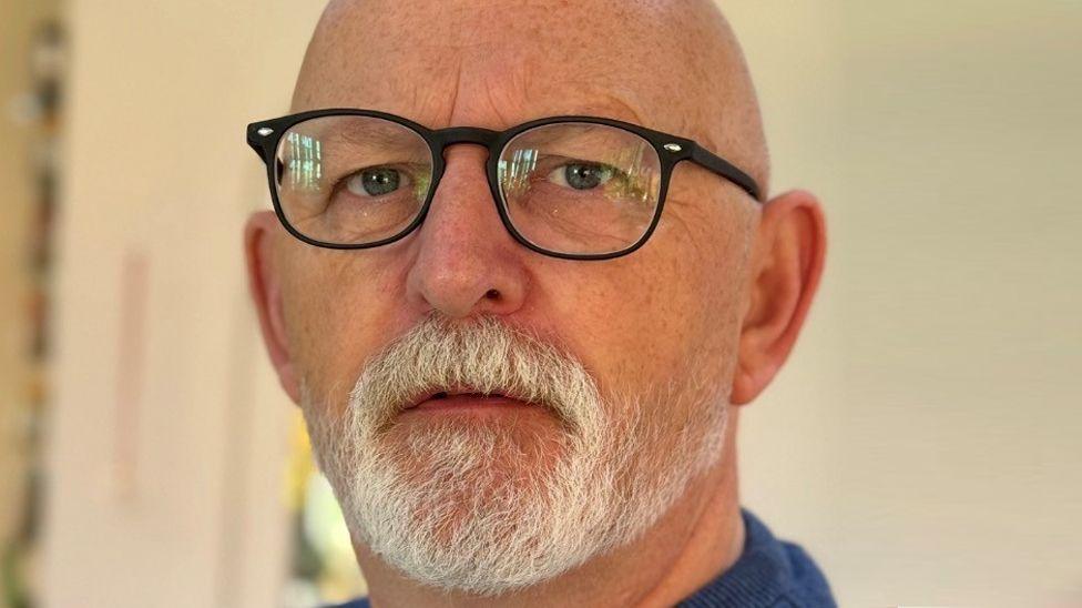 A bald man with a white beard is wearing glasses