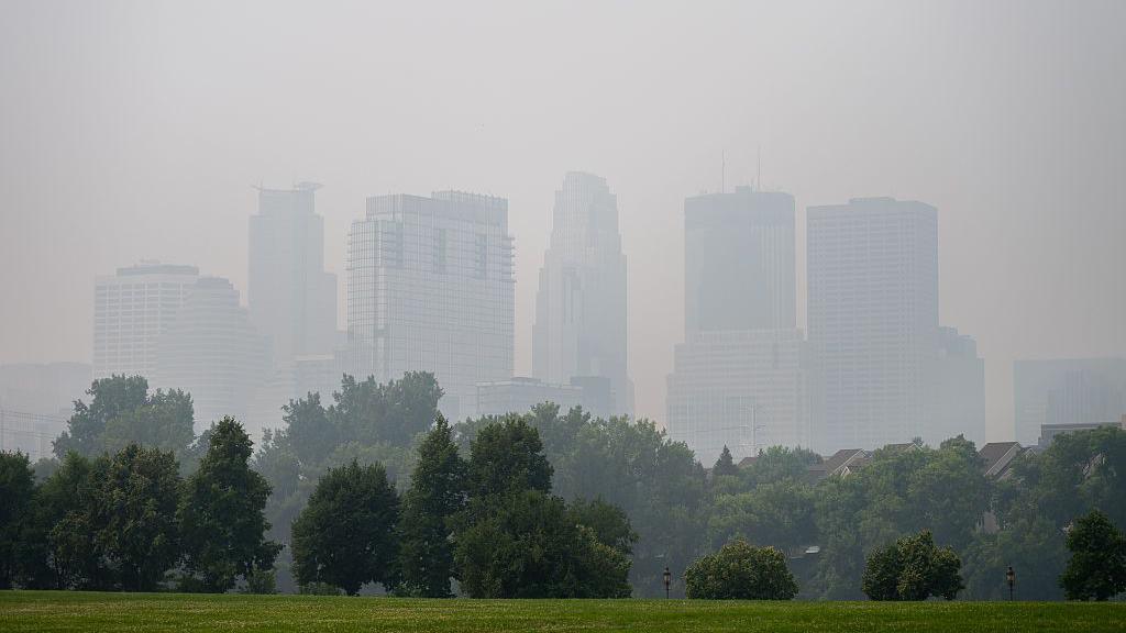 The Minneapolis, Minnesota skyline is blurred by smoke, with green park grass and trees in the foreground