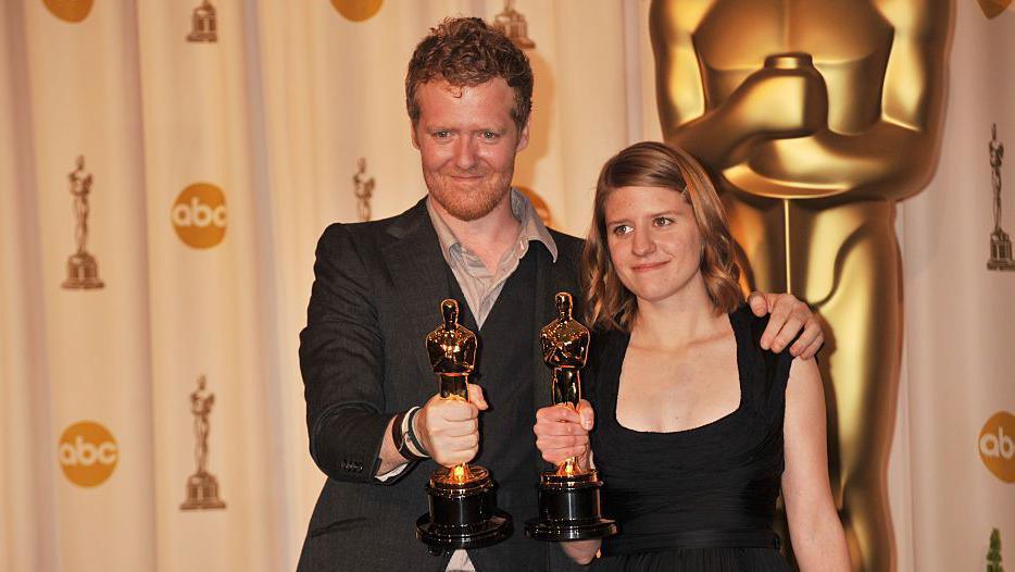 Glen Hansard and Markéta Irglova hold their Oscars. Hansard has short red hair and stubble and is wearing a suit jacket and open-necked shirt. Irglova is wearing a black dress and has shoulder-length reddish/brown hair