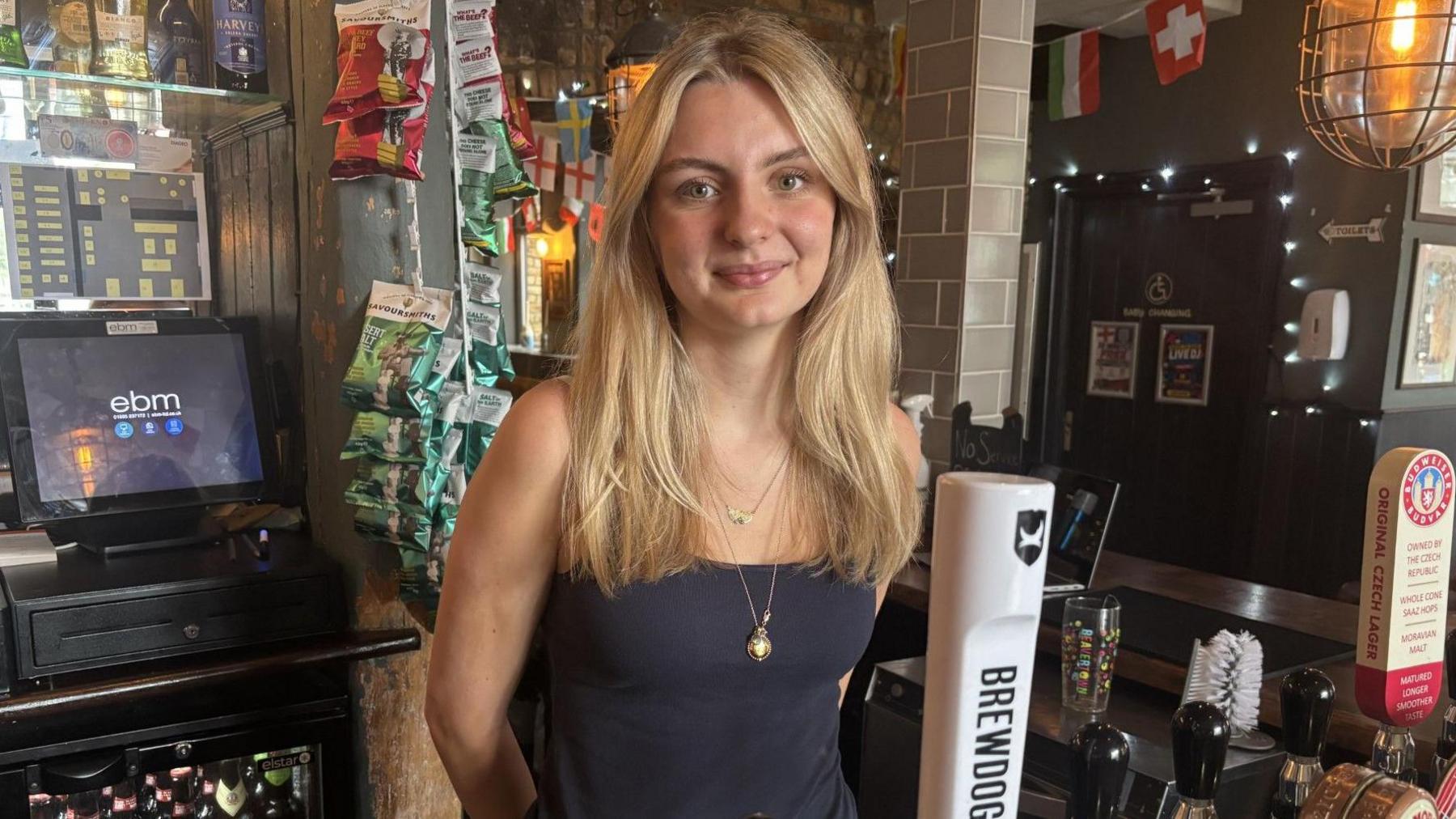 A blonde young woman stood behind a bar.