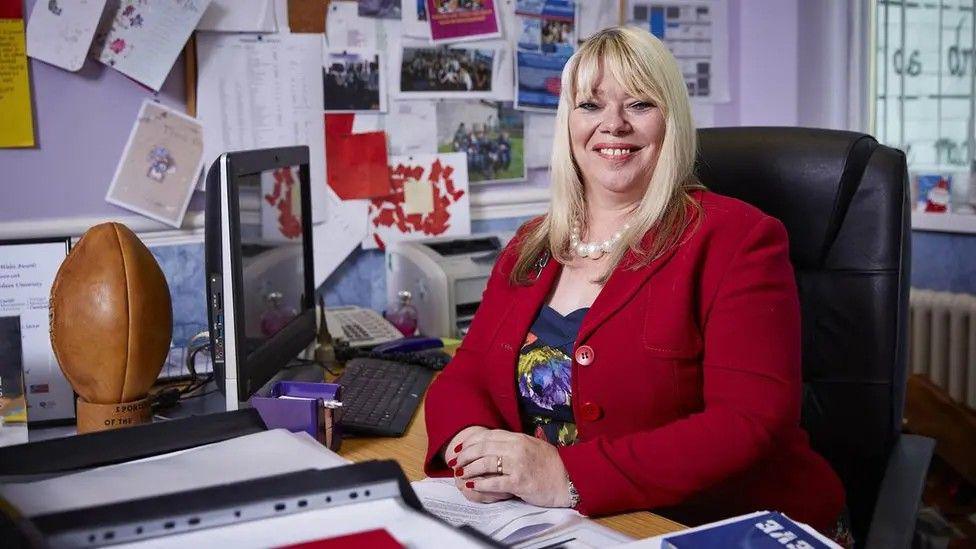 Joy Ballard a blonde haired woman, sat at her desk and smiling at the camera, wearing a red jacket and dress.