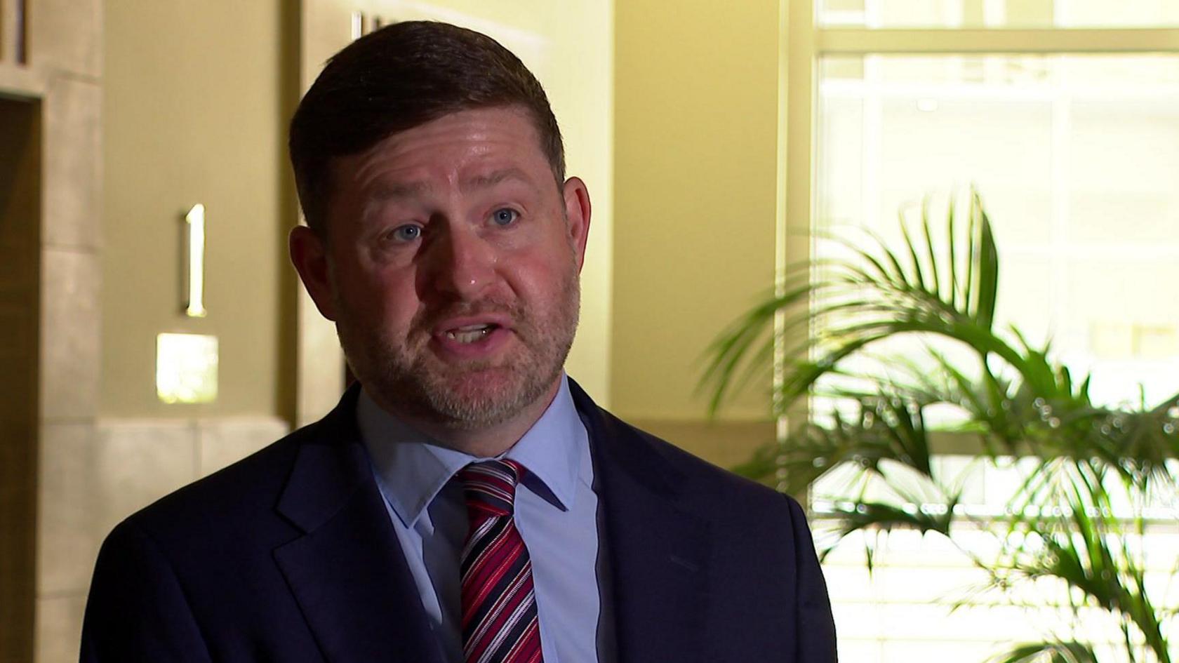 A middle aged man with short light brown hair and stubble, wearing a dark blue suit, speaks to the camera with a serious expression.