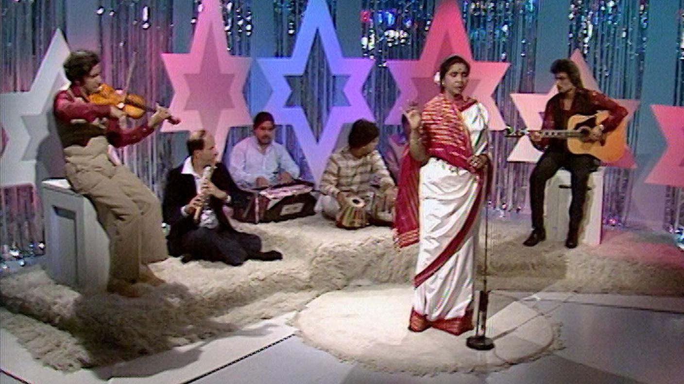 A woman in a white and red sari singing into a microphone while musicians are sat behind her playing instruments. The stage is decorated with large colourful stars and shiny curtains.