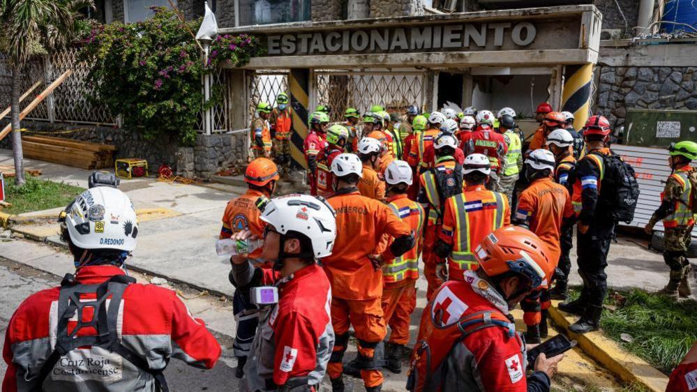Rescue workers from Mexico, El Salvador, and Costa Rica are standing in front of the entrance to the site of a collapsed multi-storey car park which collapsed  in Catia La Mar. They are wearing helmets and their uniforms. The word "estacionamiento" (Spanish for car park) can be seen on the remains of the entrance.