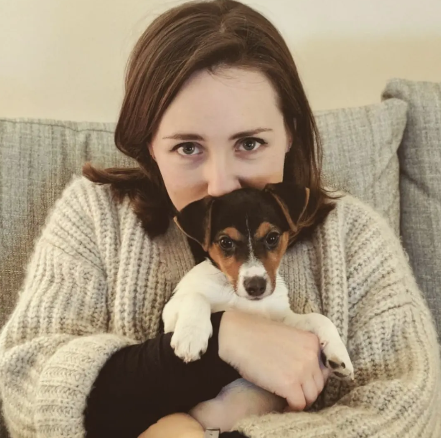 Person in a cream knitted sweater sitting on a grey sofa, holding a small black, brown, and white puppy close to their chest. The puppy faces the camera with its front paws resting over the person's arm, while a neutral-colored wall and sofa cushions form the background.