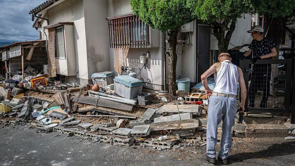 A man in a singlet and blue jeans surveys the debris outside a house. Another person in a cap and gloves looks on from behind a fence