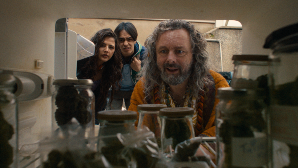 A man with long, curly grey hair and facial hair, staring into a cupboard full of jars. A young man and young woman, both brunette, are stood behind him, also leaning down to look at what he is looking at. They have puzzled looks on their faces.