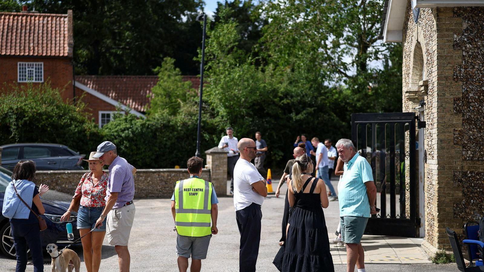 Evacuated residents gather at Westleton Village Hall, in Westleton. It is a sunny day. One person has a dog. Groups are talking to each other outside the hall.