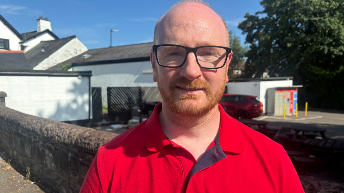 Sion is looking at the camera wearing a red polo top and glasses. Behind him is a small car park and parcel lockers