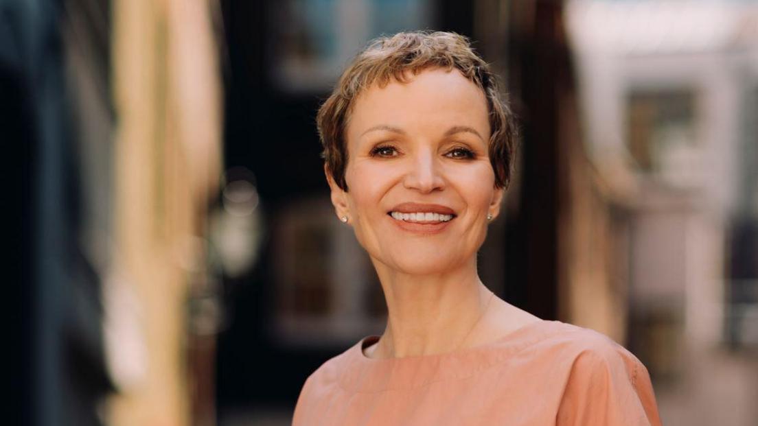 Marcia Kilgore with cropped short brown hair smiling at the camera with a blurred background wearing a nude coloured round neck top