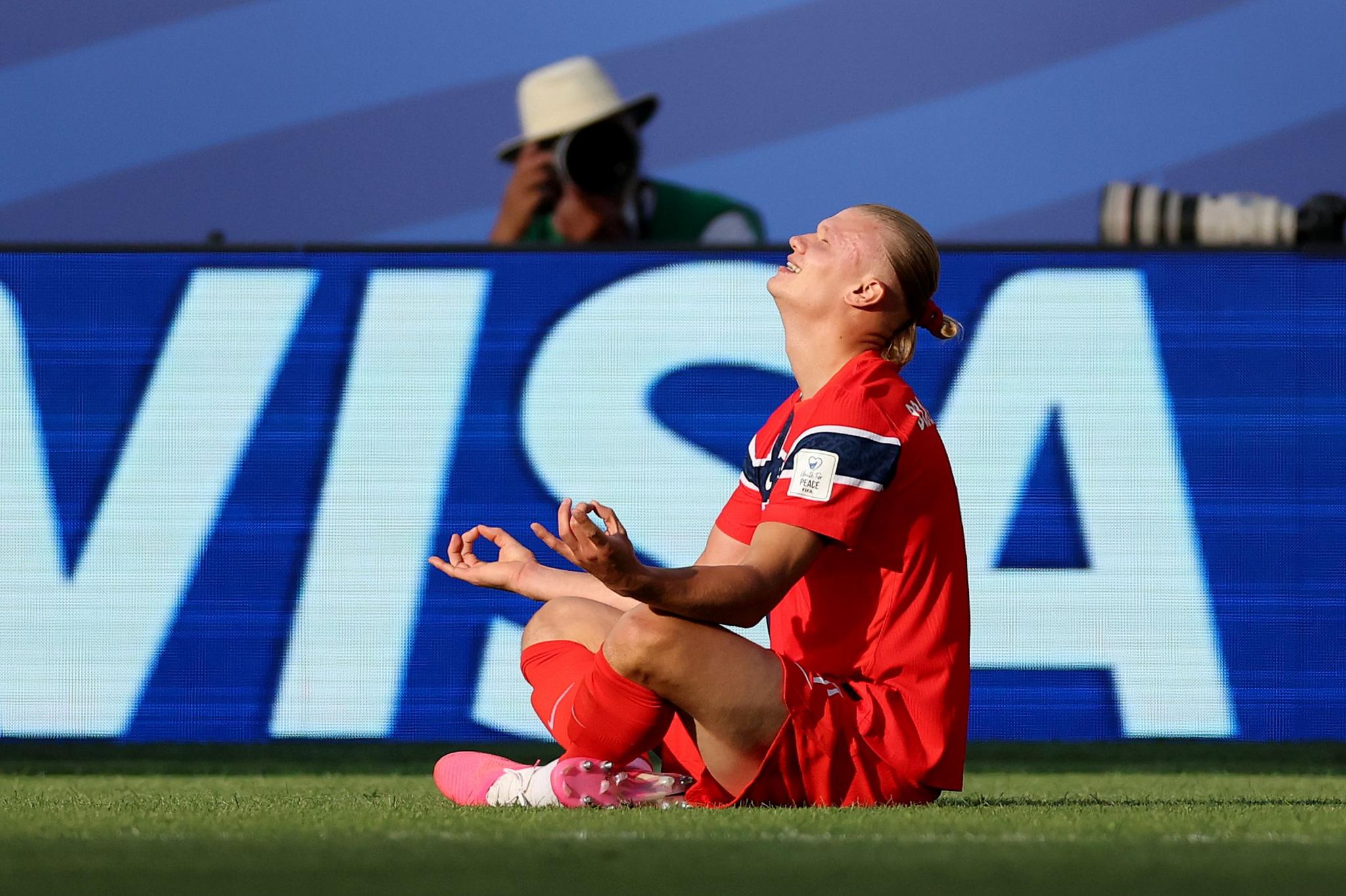 Haaland sits cross legged on the ground with his hands rested atop his knees. His eyes are closed and he has a wide smile.