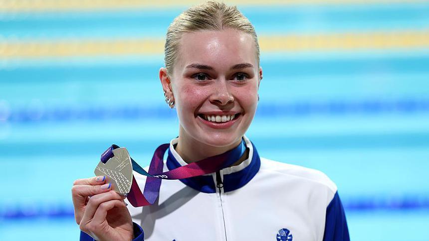 The swimmer Katie Shanahan holding her silver Commonwealth Games medal in front of the pool. She has blond hair, is grinning with delight and is wearing a white and blue athletic/tracksuit top.