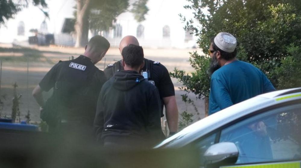 Two male police officers and two other men in the grounds of Shrubland Hall at Barham, Suffolk. The officers are in the middle of the pic, one with his back the head of a third man, who is dressed in black. On the right is a man with a long beard dressed in blue Islamic dress and a white cap. A police car is in the right foreground, side on and showing its side window only.