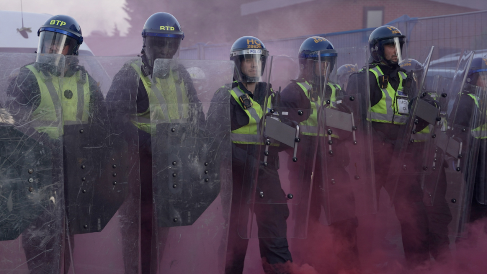 Police officers stood with shields surrounded by pink dust amid clashes with protesters