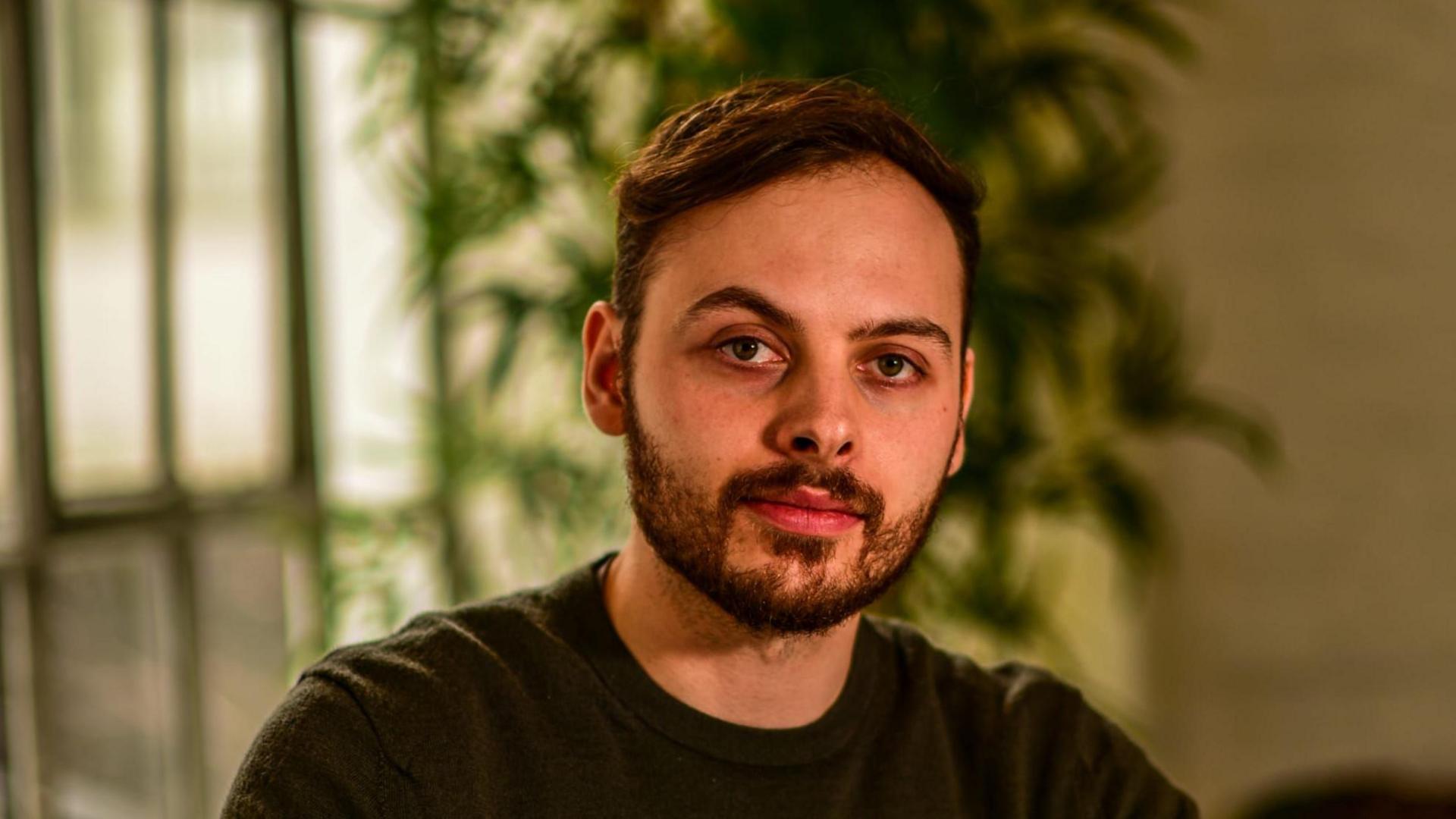 Rees Cowne looking at the camera while sat in a room, with a green plant behind him blurred out. He has short brown hair and stubble and is wearing a brown top.