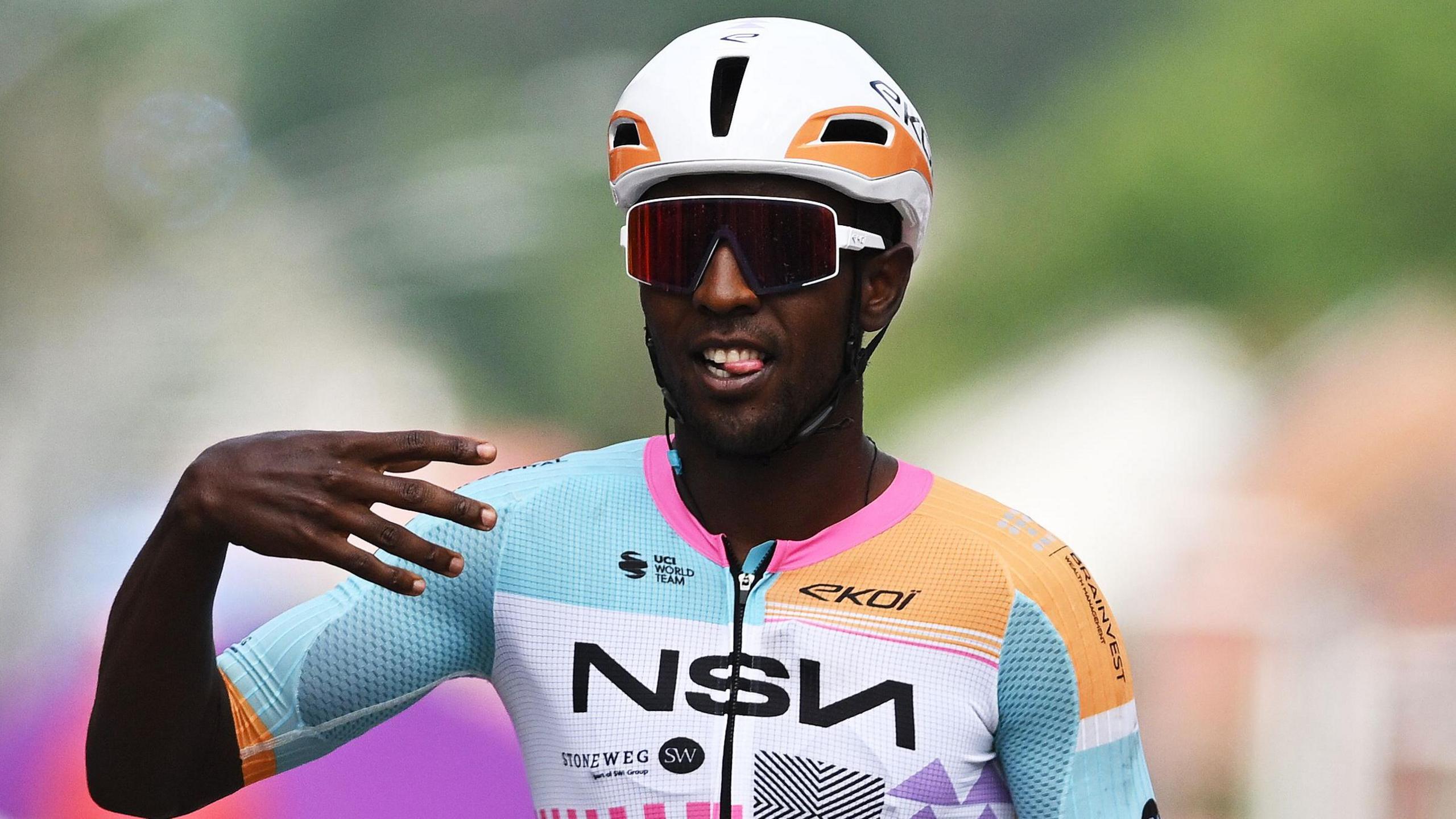 Biniam Girmay, visible from chest up, rides while wearing a white cycling helmet, sunglasses and a pink, orange, light blue and white cycling top. He raises his right hand, bent at the wrist and with fingers spread, as he bites his tongue in celebration