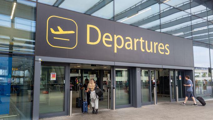 The departures entrance at Gatwick Airport with large yellow "Departures" signage above glass doors. Travellers with luggage can be seen entering the terminal building.