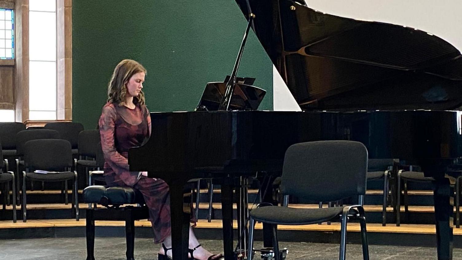 Freya Terris sits at a grand piano, wearing a long sleeved dark dress with red flowers, and black strappy heels. The room behind her has green walls and large, long windows. She is also surrounded by chairs.