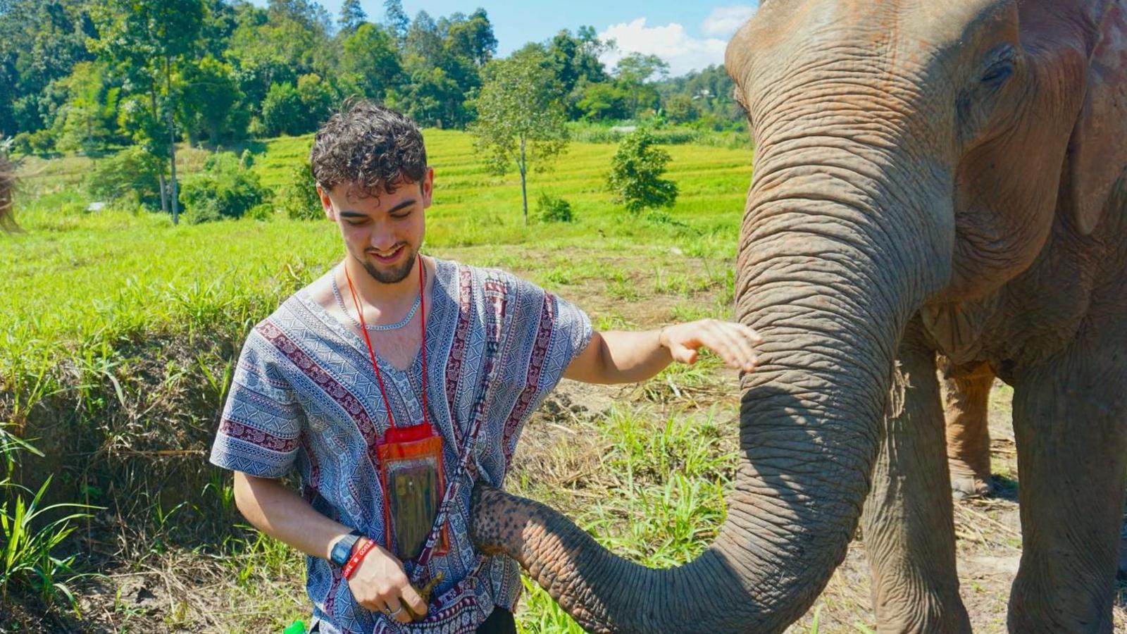 Calum Macdonald pets an elephant as it touches his striped patterned shirt with its trunk. Behind them are green paddy fields and tropical woods beyond that. He has short black curly hair.