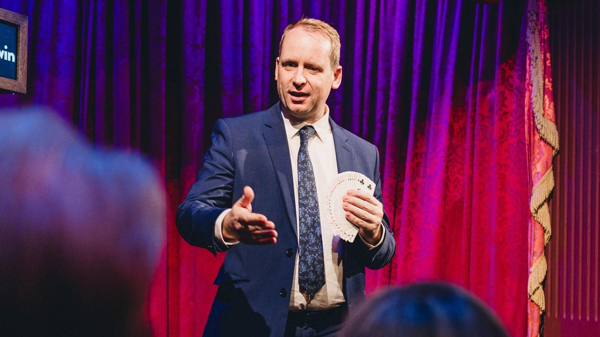 A man with short blonde hair stands on a stage holding a fanned out pack of cards. There are thick red curtains behind him, and some of the audience can be seen from behind. He is wearing a navy suit and a dark blue floral tie.