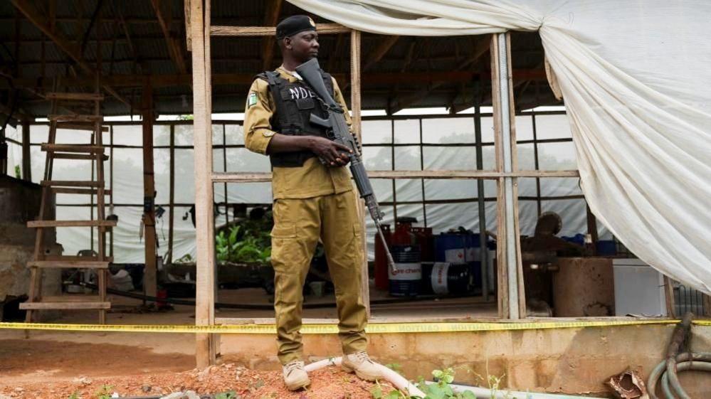 An armed officer in khaki uniform and black stab vest stands guard in front of a makeshift room.