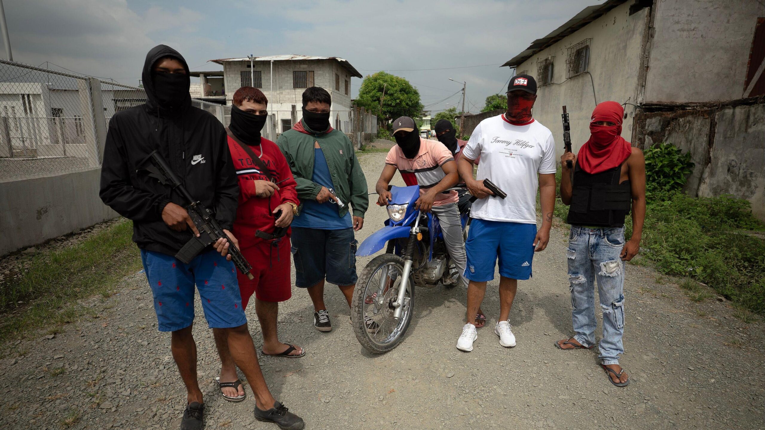 Seven young men in balaclavas brandish automatic weaponry in broad daylight, facing the camera. two of the men are on a dirt bike.