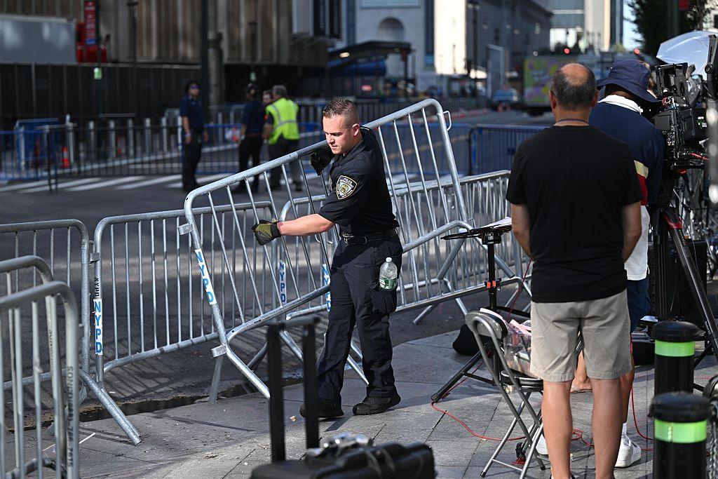 A police officer places a metal barricade on the streets around Madison Square Garden while a TV crew looks on.