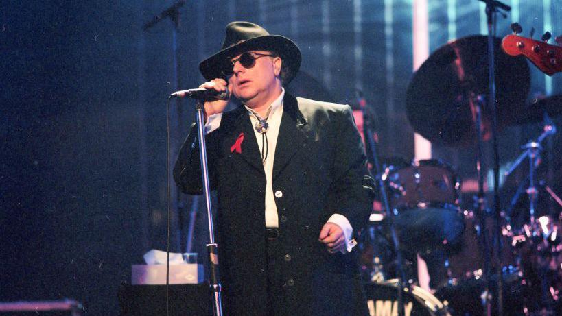 Van Morrison is dressed in black and is wearing a black hat and shades while singing on stage