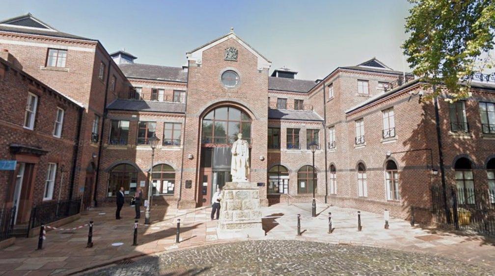 Street view of Carlisle Crown Court. To the right in the foreground there is part of a green tree. In the centre there is a plinth with a yellowing statue. In the background there is a red brick building with large glass windows. At the top of the building there is a crest.