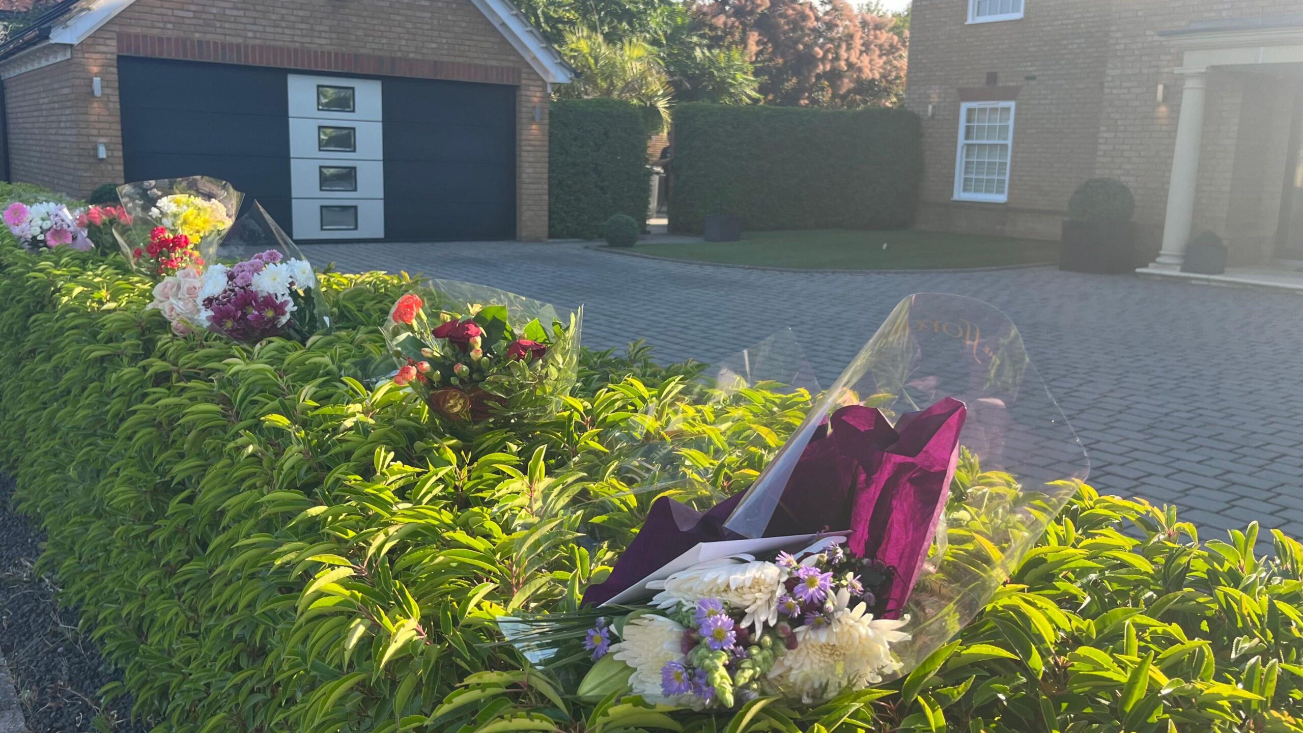 Bunches of plastic-wrapped flowers pushed into a green hedge in front of a house in Carnoustie Drive in Great Denham. Behind the hedge is a garage and a corner of the detached brick-built house and its front entrance.