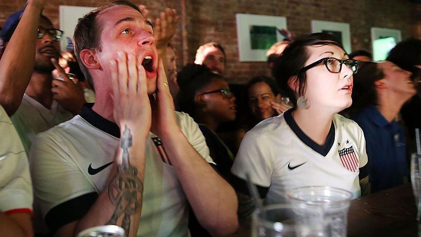Two United States football fans react during the match between Ghana and USA at the WoodWork bar in the Brooklyn borough of New York City.  One is a man and the other is a woman. They are both wearing USA football tops.