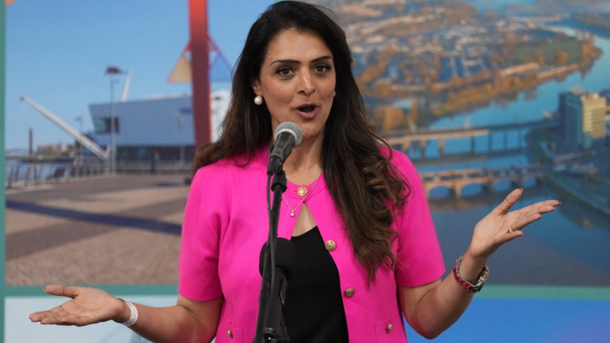 Natasha Asghar speaking into a microphone mounted on a stand. She is wearing a bright pink short-sleeved jacket with gold-coloured buttons over a black top. Pearl-style earrings, a necklace, a wristwatch and bracelets are visible. Both hands are raised and held out to either side in a gesturing pose while speaking.