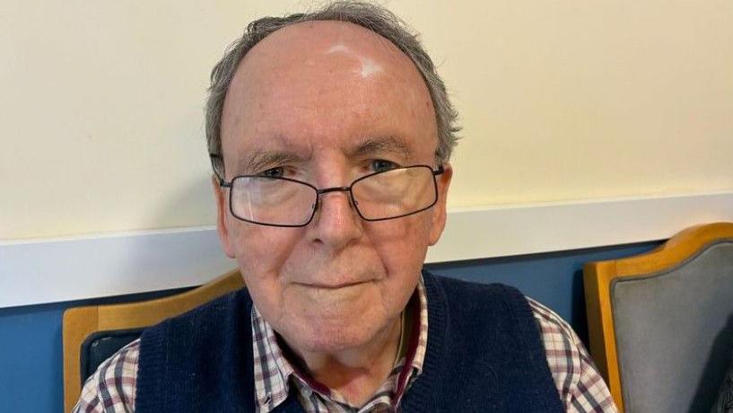 Richard is an elderly gentleman smiling slightly at the camera. He has lost most of his hair and wears thick spectacles with a navy tank top over a white, burgundy and navy chequered shirt. He is sitting on a wooden chair with a blue leather backrest, up against a cream wall.