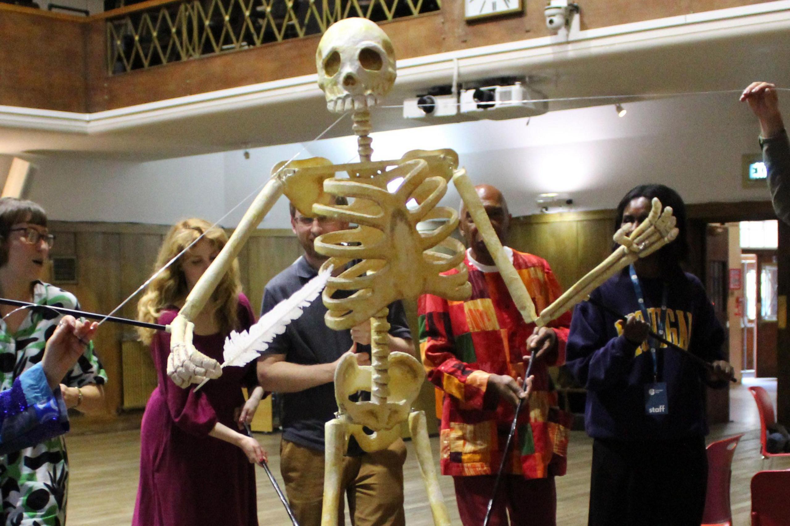 A group of five people animate a giant puppet depicting the skeleton of Thomas Paine