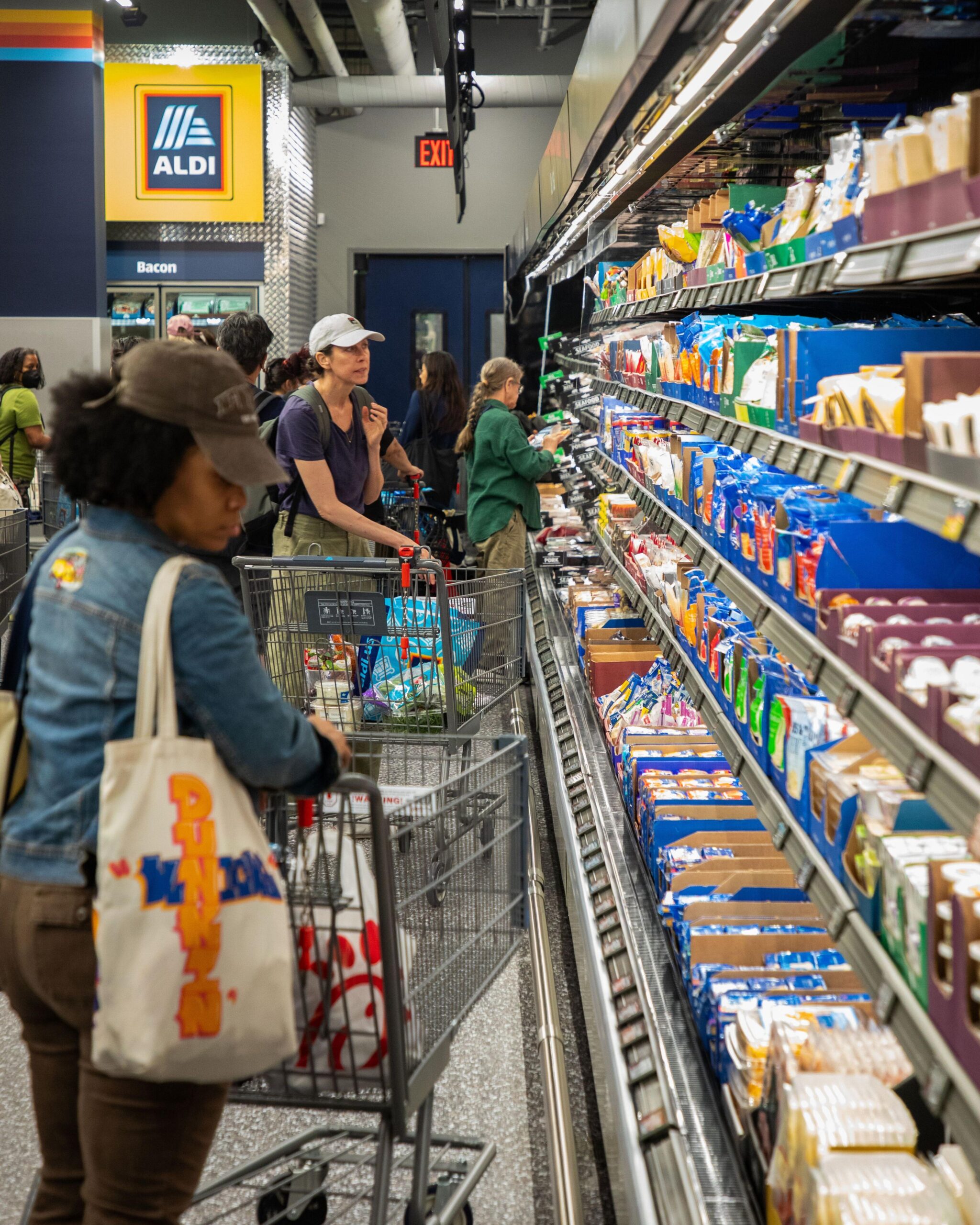 A woman in a jean shirt with brown jeans and a brown hat looks at cheese options while holding onto her shopping cart. Another woman in the background wearing green pants, a blue shirt and a beige hat looks at other dairy options, including a third woman in a green shirt, khaki pants looks at the labeling of an item.