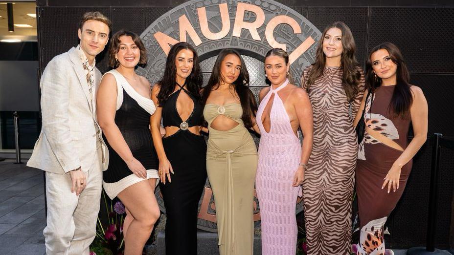 A man on the left stands next to six women in front of a sign saying 'MURCI' . He wears a cream linen suit, while the women all wear maxi dresses of different colours and patterns.