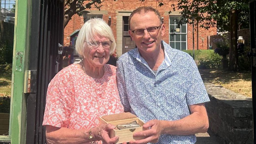 Standing in front of green gates, a tall and slender Filip Krapels wearing a short sleeve blue shirt, hands the cigarette case lid to Diane Owen, an elderly lady with white hair and glasses. Each has one hand supporting the display box in which it was transported