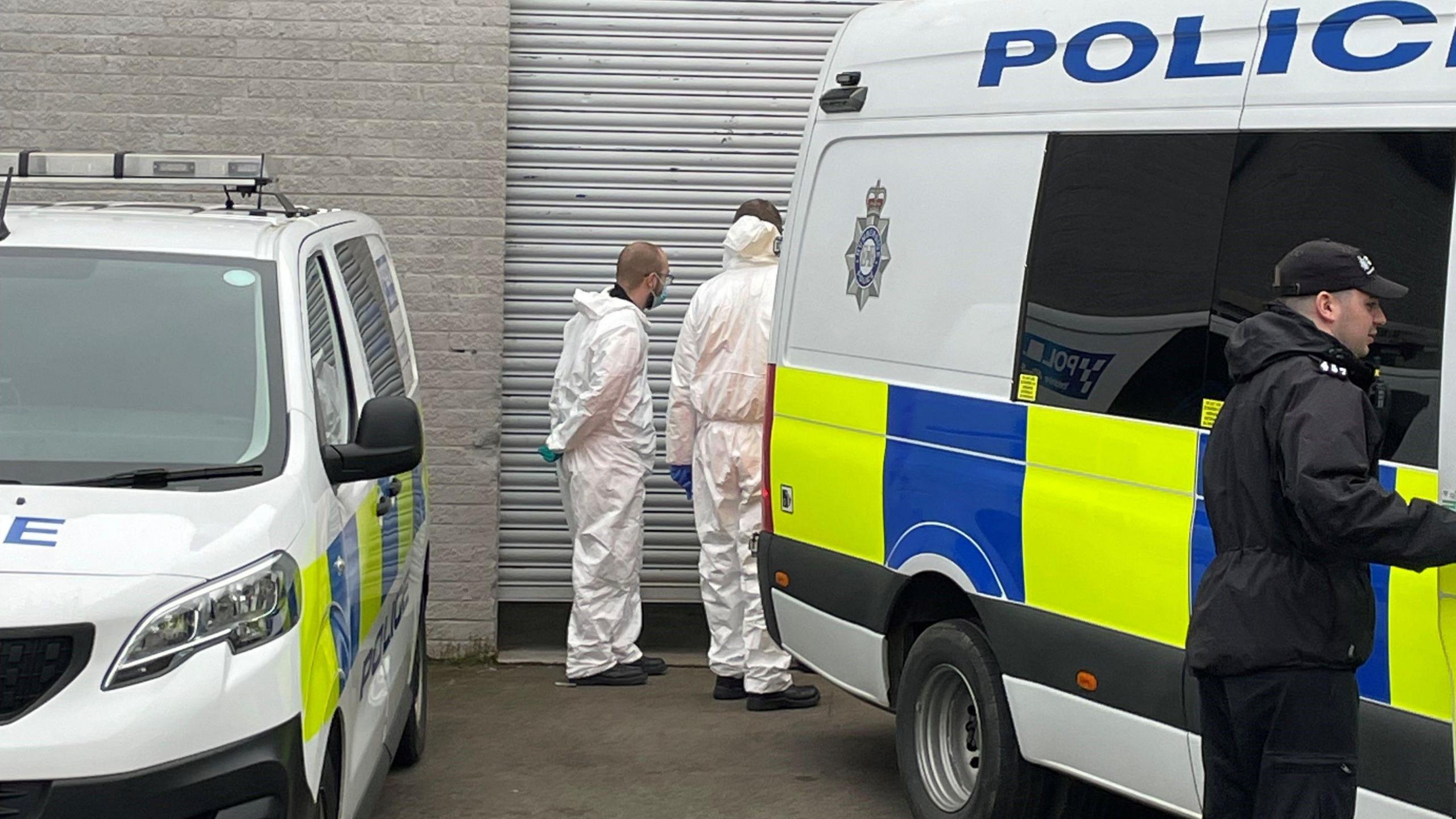 Two people in white forensic suits stand in front of a metal door. To each side of them is a police vehicle.