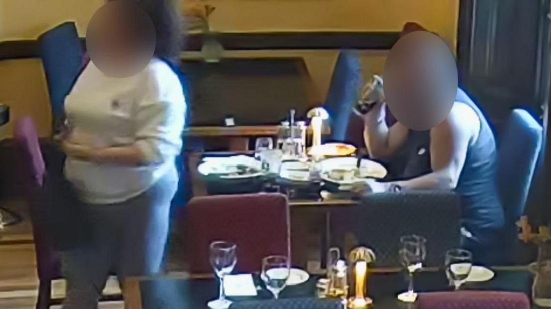 A man in a vest sat at a table with empty plates  in front of him. He is drinking from a pint glass of coke. His face is blurred. On the other side of the table, a woman in grey leggings and a white sweatshirt is stood. Her face is also blurred. There are other restaurant tbales set with plates, cutlery and glasses around them.