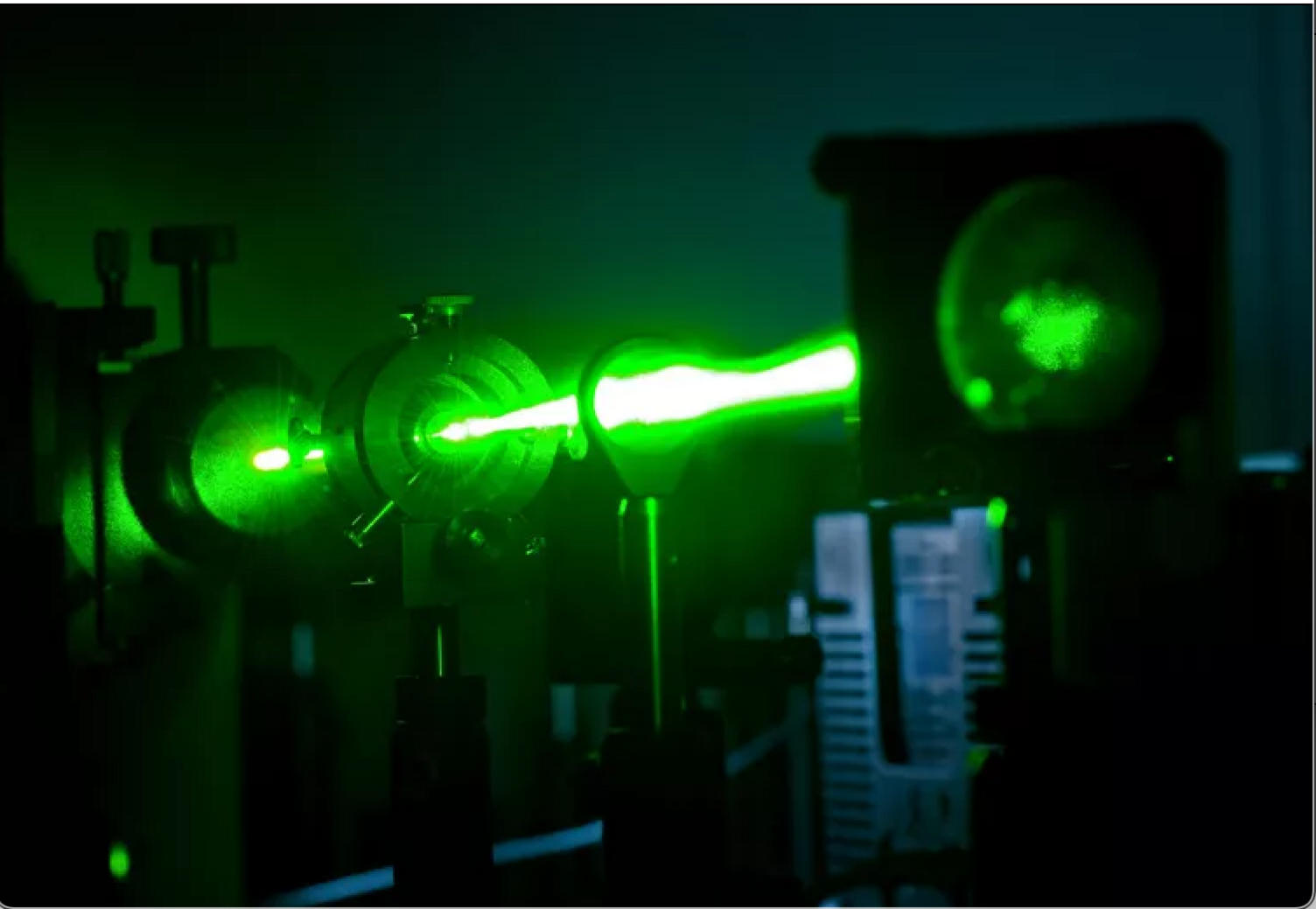 A bright, glowing green laser beam shoots horizontally through a dark laboratory. It passes through circular metal instruments and lenses, getting wider and more intense. The surrounding equipment is mostly in shadow, so the vivid green light is the dominant, almost otherworldly feature against the black background.