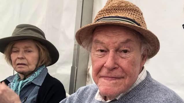 A close-up selfie-style photo of two people seated side by side in an  outdoor event space. In the centre, Timothy West is sitting slightly behind the camera holder, wearing a woven brown hat and a light grey sweater over a collared shirt. On the left, Prunella Scales is partially in frame, seated further back. She is wearing a darker hat, a scarf, and a jacket, and is positioned slightly turned away from the camera.