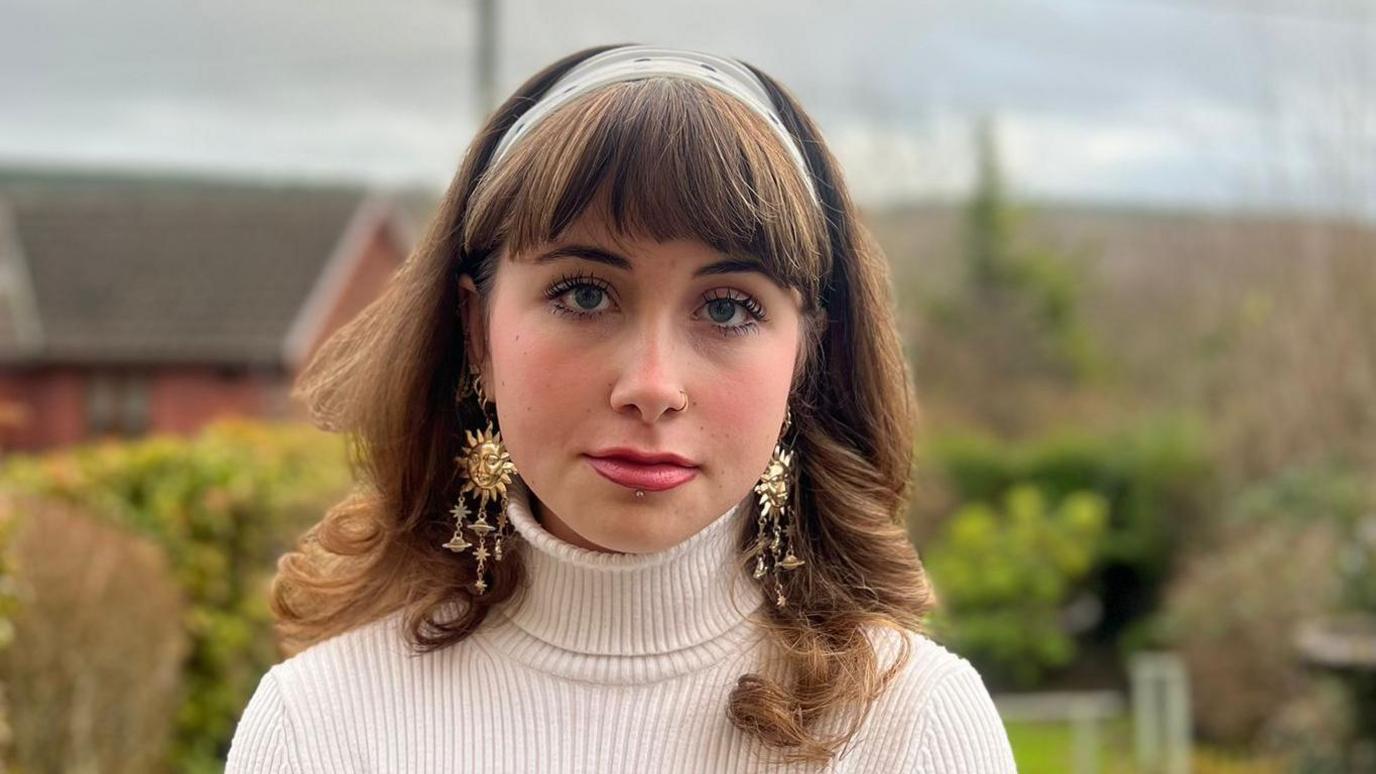 Maddie is stood outside in a back garden. She has light brown hair and fringe. She is wearing a polkadot hair band, white knitted turtleneck jumper, and large gold earrings. She has her nose pierced on the right side of her face and a stud piercing under her bottom lip.