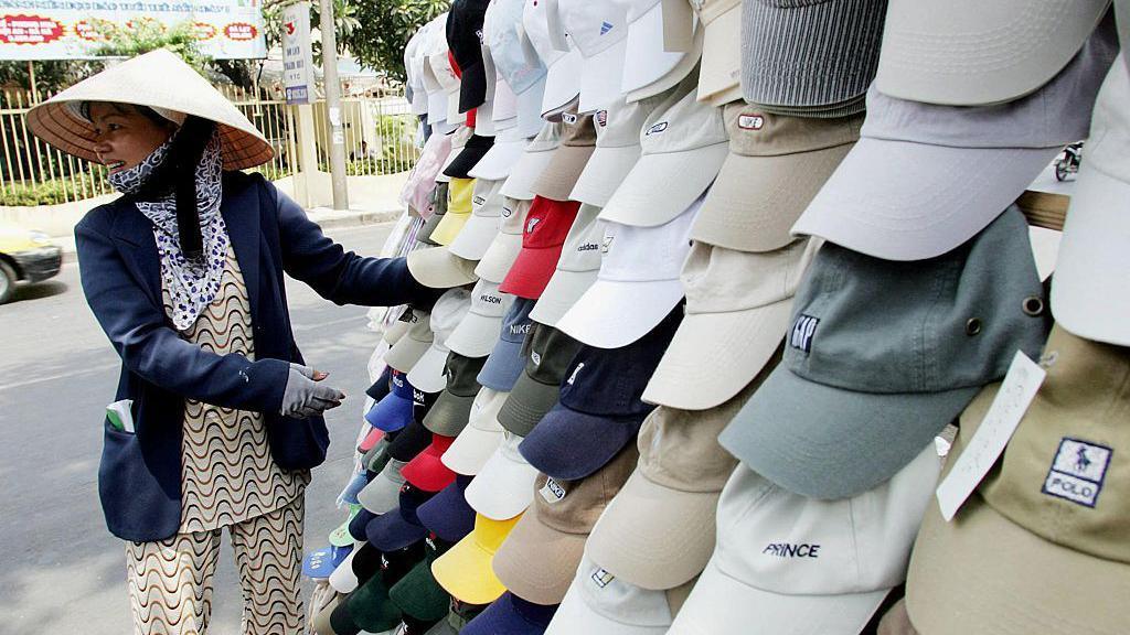 Caps bearing the logos of Polo, GAP, Prince and others hang in the foreground next to a woman wearing a blue blazer and a straw nón lá hat