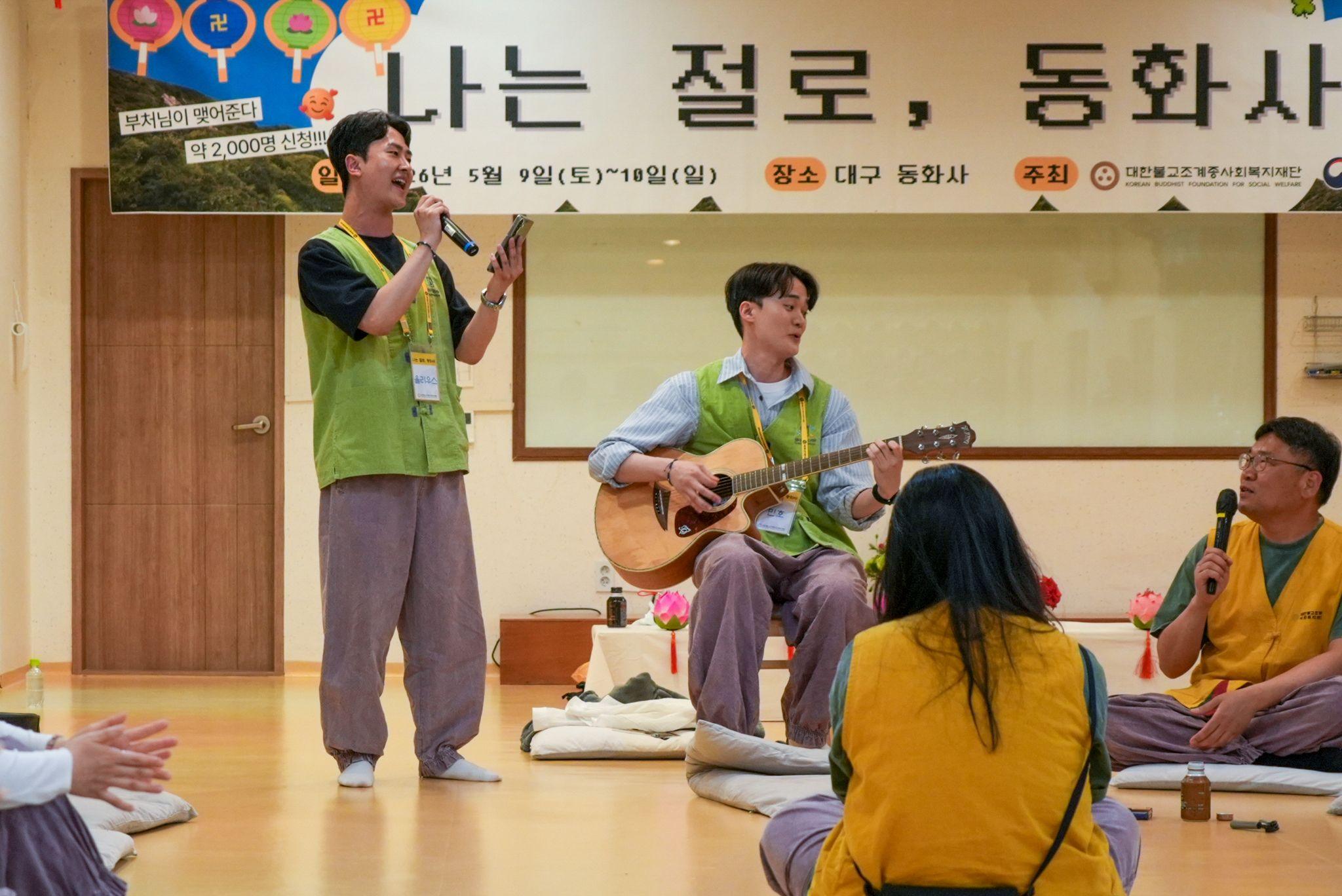 Participants sit and perform music at the temple dating retreat - one person stands holding a microphone, while another sits playing an acoustic guitar. Other attendees sit on floor cushions in a circle. A large banner with Korean text hangs behind the group.