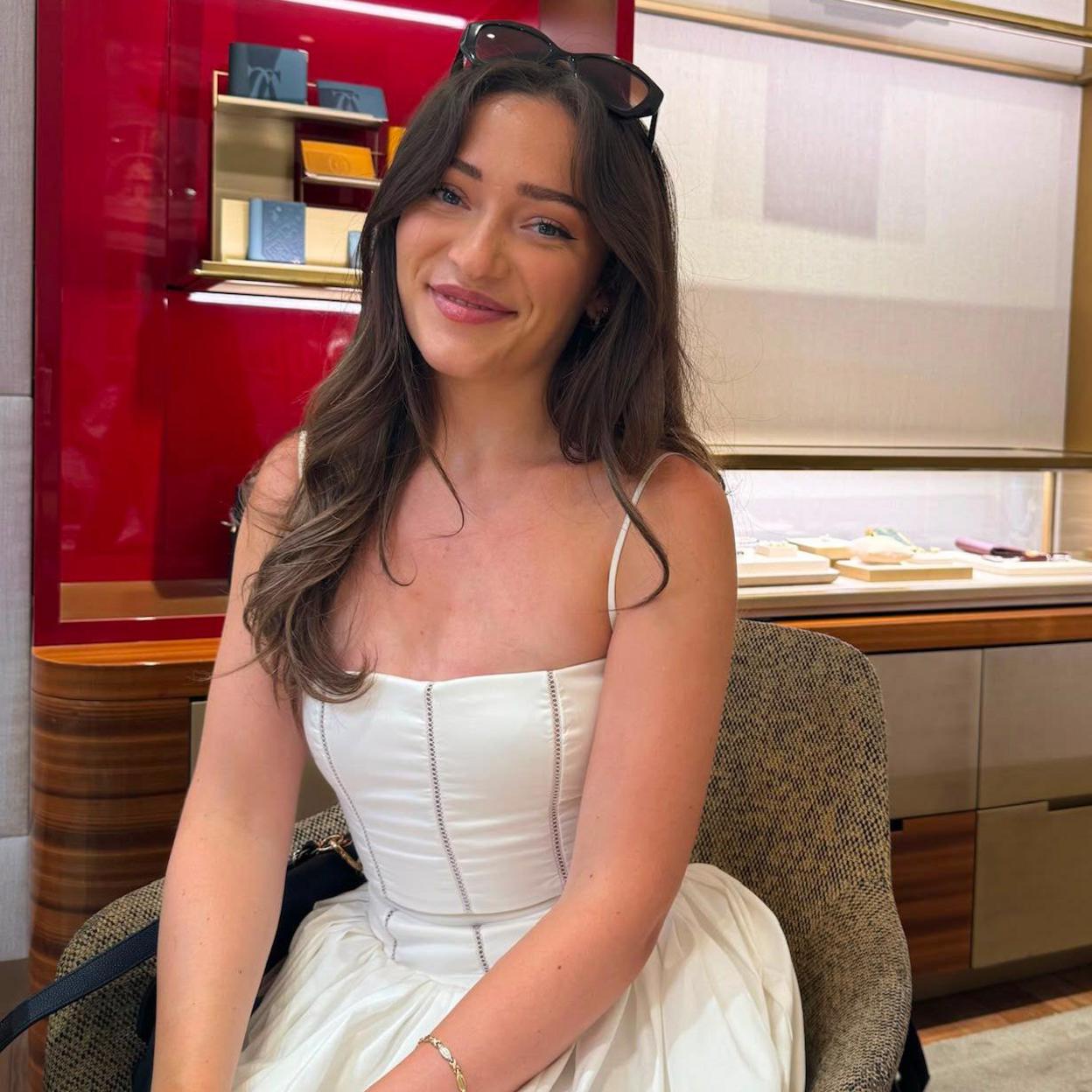 Taya Reed has long dark hair and smiles at the camera, sitting perched on a chair, wearing a white dress