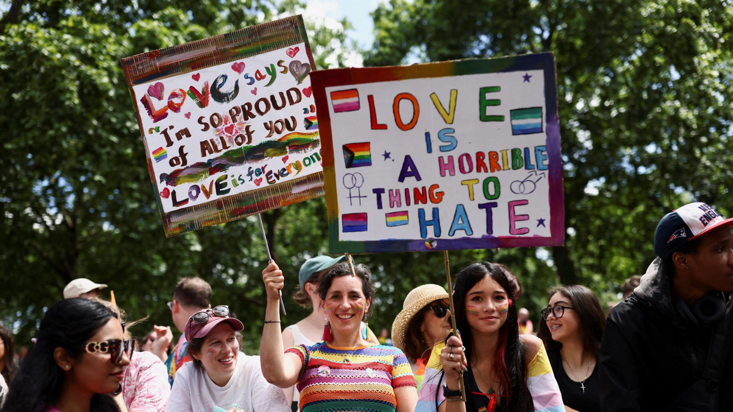 A crowd of smiling attendees at a Pride event under a canopy of green trees. In the centre, two women hold up colorful, hand-painted protest signs; the sign on the left reads "Love says I'm so proud of all of you," while the larger sign on the right reads "Love is a horrible thing to hate" alongside illustrations of various Pride flags.
