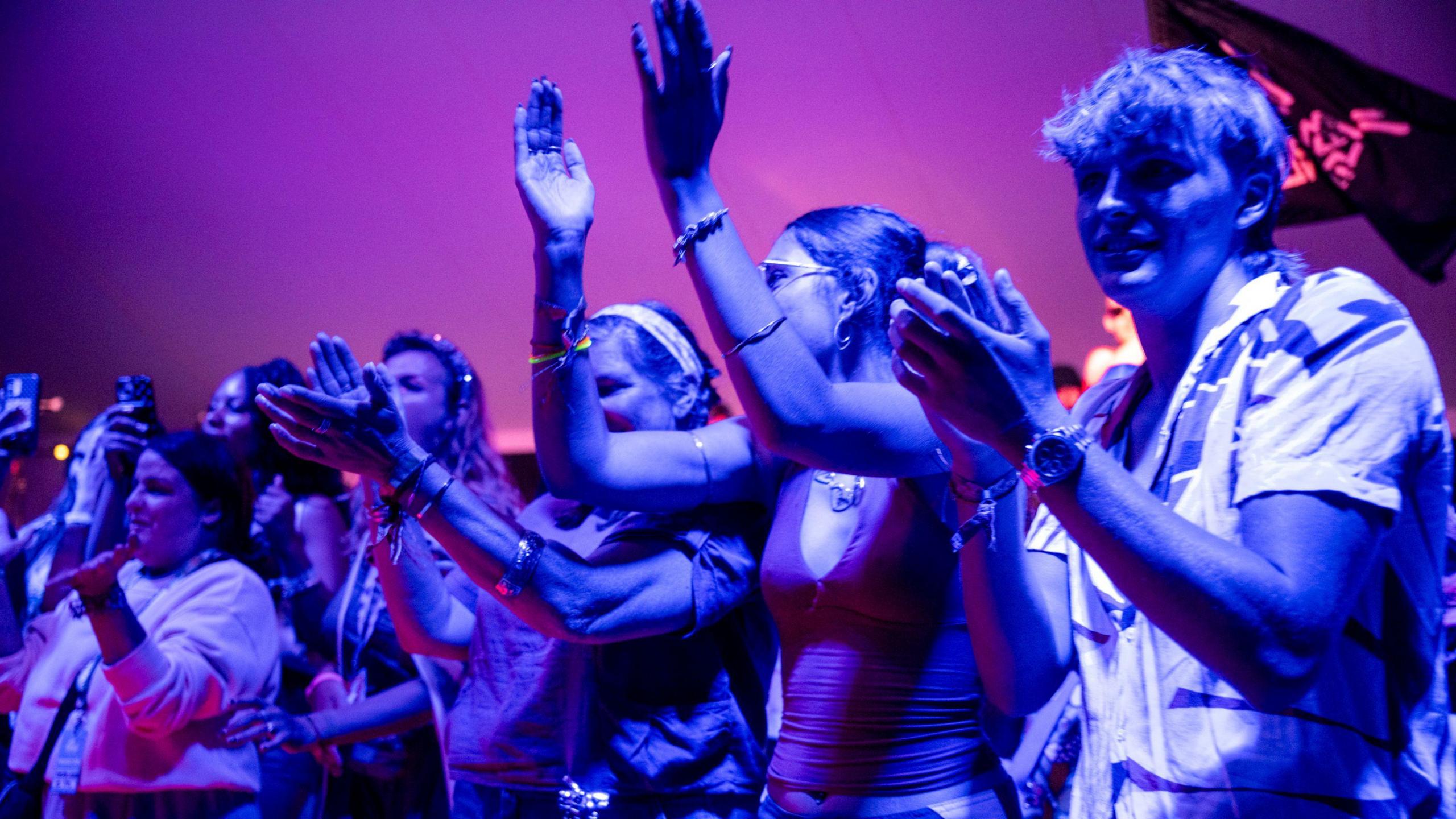 A group of several young people, illuminated by blueish light, applaud as they watch a live performance at WOMAD 2026