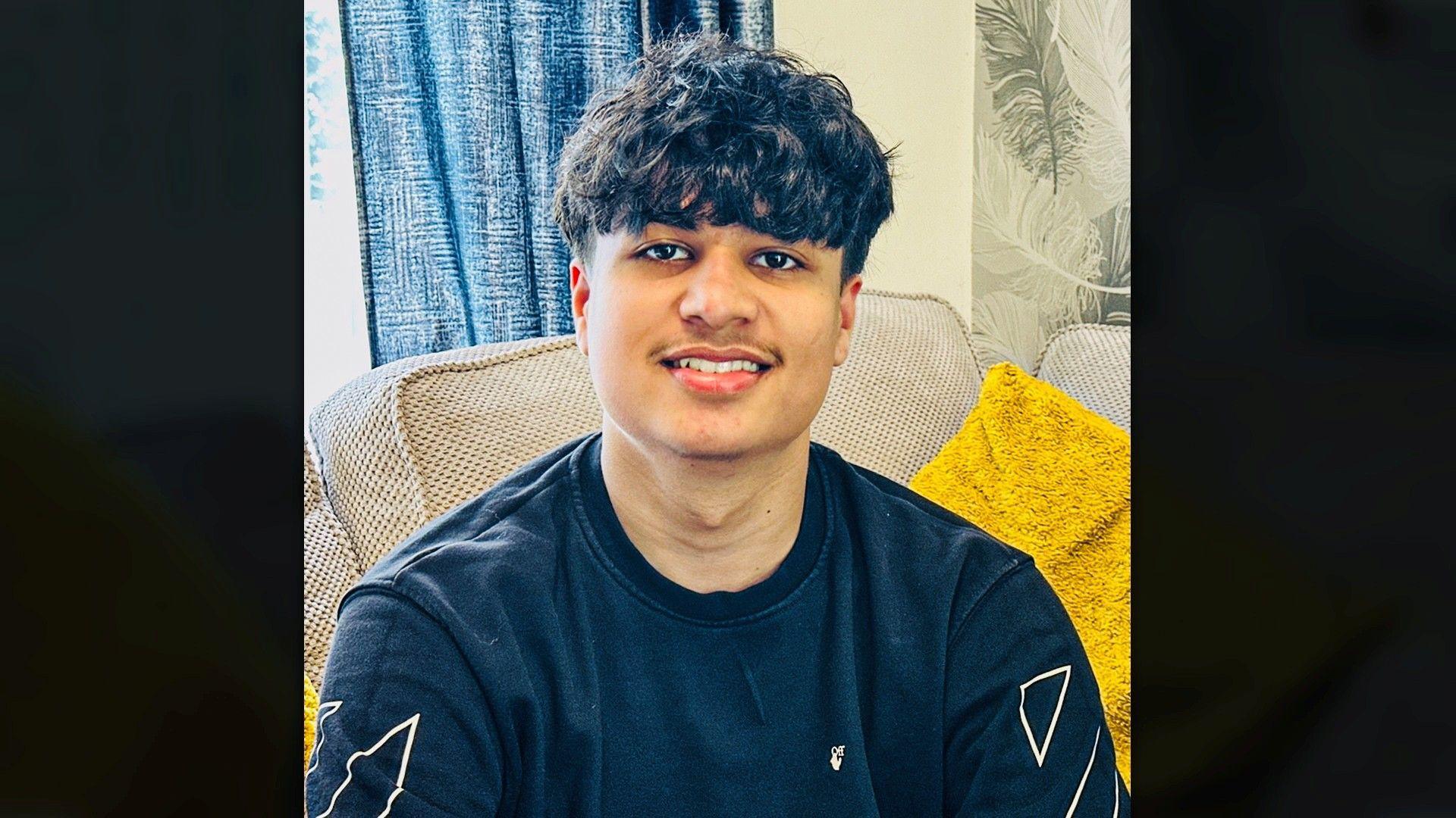 A young man sat on a beige sofa with a yellow cushion beside them. He is wearing a dark sweatshirt with white geometric designs, with a blue curtain and leaf‑patterned wallpaper behind them.