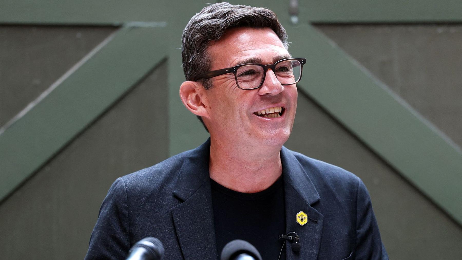 Andy Burnham smiling and wearing a black suit in front of a green door
