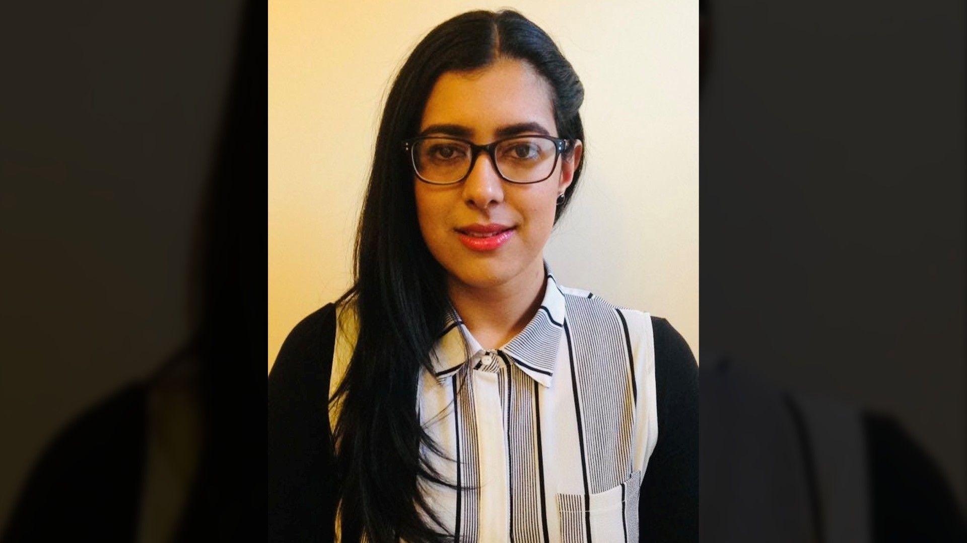 A woman with long dark hair is shown in a simple portrait against a plain light‑coloured wall. She is wearing glasses and a black‑and‑white striped collared shirt.